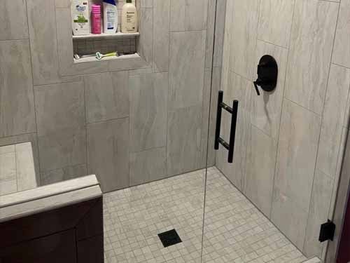 Custom tile shower with large format gray tile, mosaic floor, built-in niche, and frameless glass door