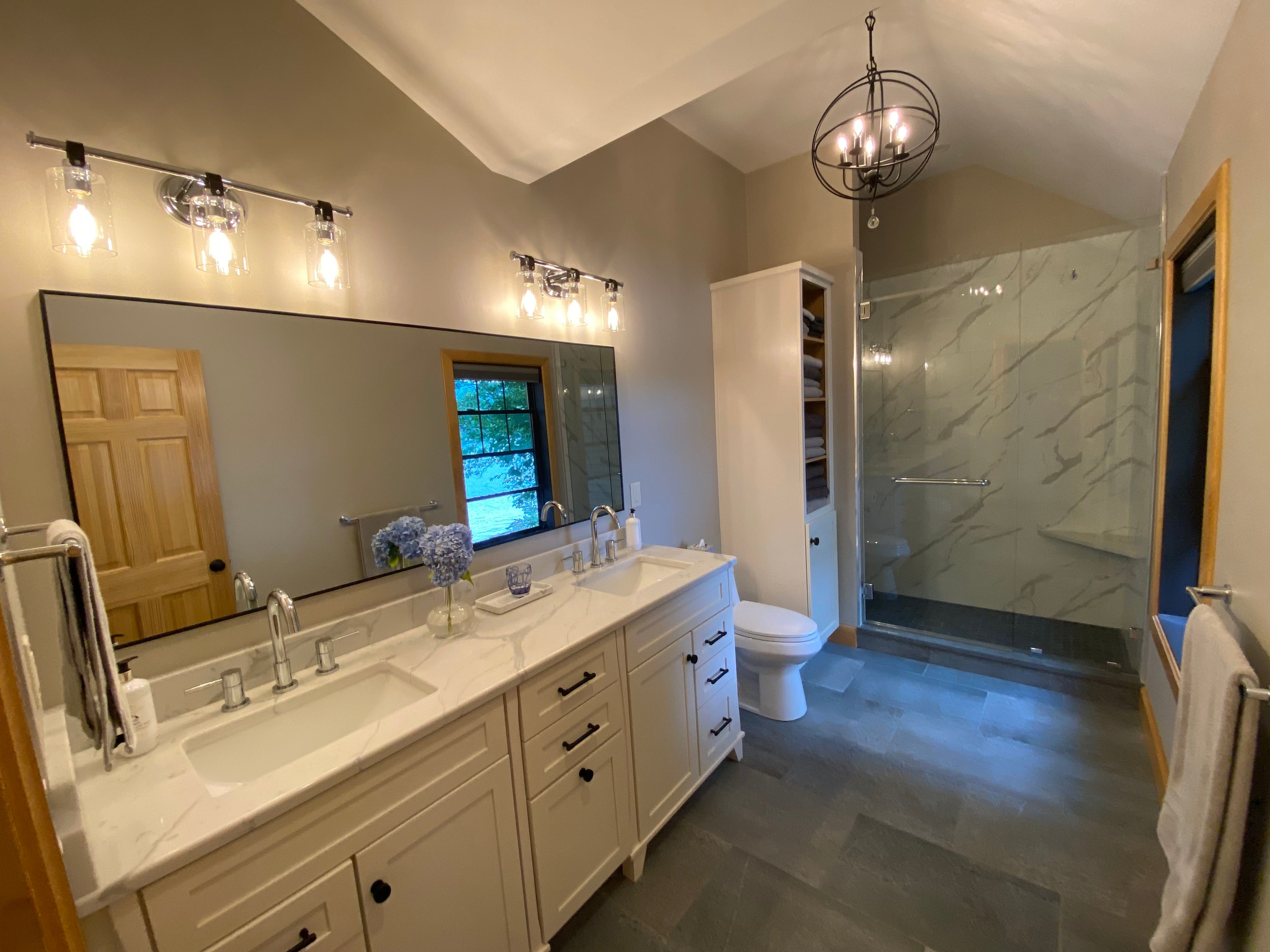 Spacious master bath with white double vanity, quartz countertop, orb chandelier, and marble tile shower