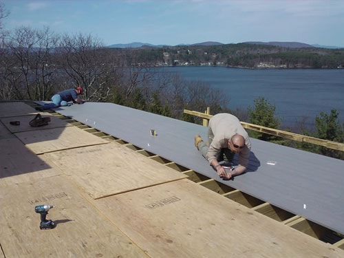Two contractors installing composite deck boards on elevated lakeside deck with lake and mountain views