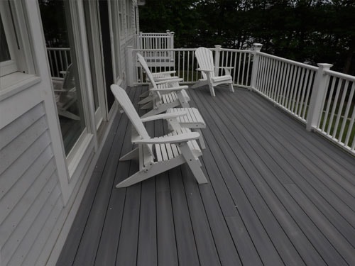 Completed gray composite deck with white Adirondack chairs and white vinyl railings overlooking wooded yard