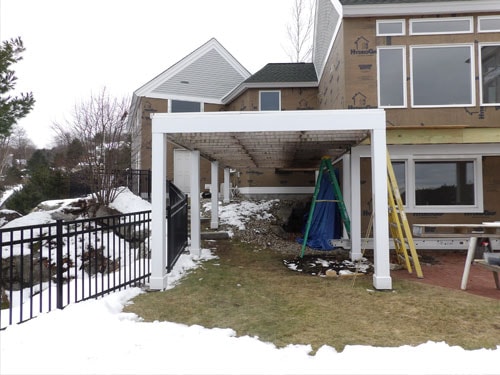 Completed white pergola structure attached to lakeside home with black iron fence and snowy yard