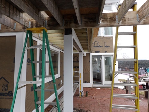 Under-deck view of pergola framing with white posts, pressure-treated beams, and brick patio below