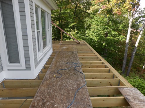 Deck framing with pressure-treated joists and AdvanTech subfloor alongside gray home with fall foliage