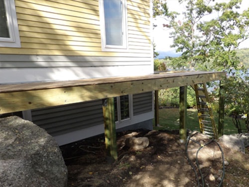 Elevated deck framing with pressure-treated posts and subfloor on yellow lakeside home with boulder landscaping
