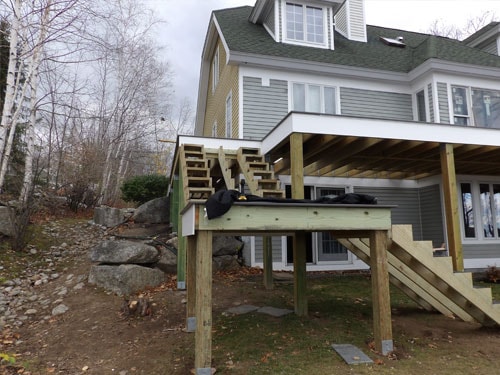 Elevated deck framing with pressure-treated posts, stair stringers, and lower platform on gray home in late fall