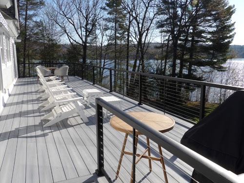 Gray composite deck with white Adirondack chairs, bar stools, Weber grill, and black cable railing overlooking lake