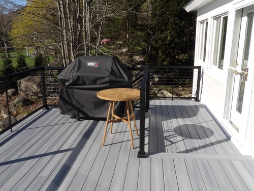 Gray composite deck with covered Weber grill, teak bar stool, and black cable railing in spring