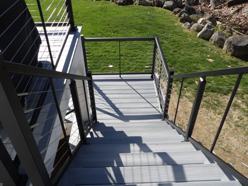 Looking down gray composite deck stairs with black cable railings and metal posts onto green lawn and stone wall