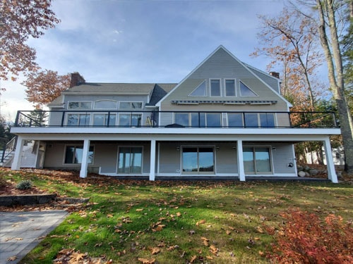 Large lakeside home rear exterior with elevated white deck, black cable railings, and upper balcony in fall foliage