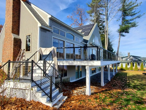 Lakeside home with elevated white deck, black cable railings, solar panels, and black metal stairs in fall foliage