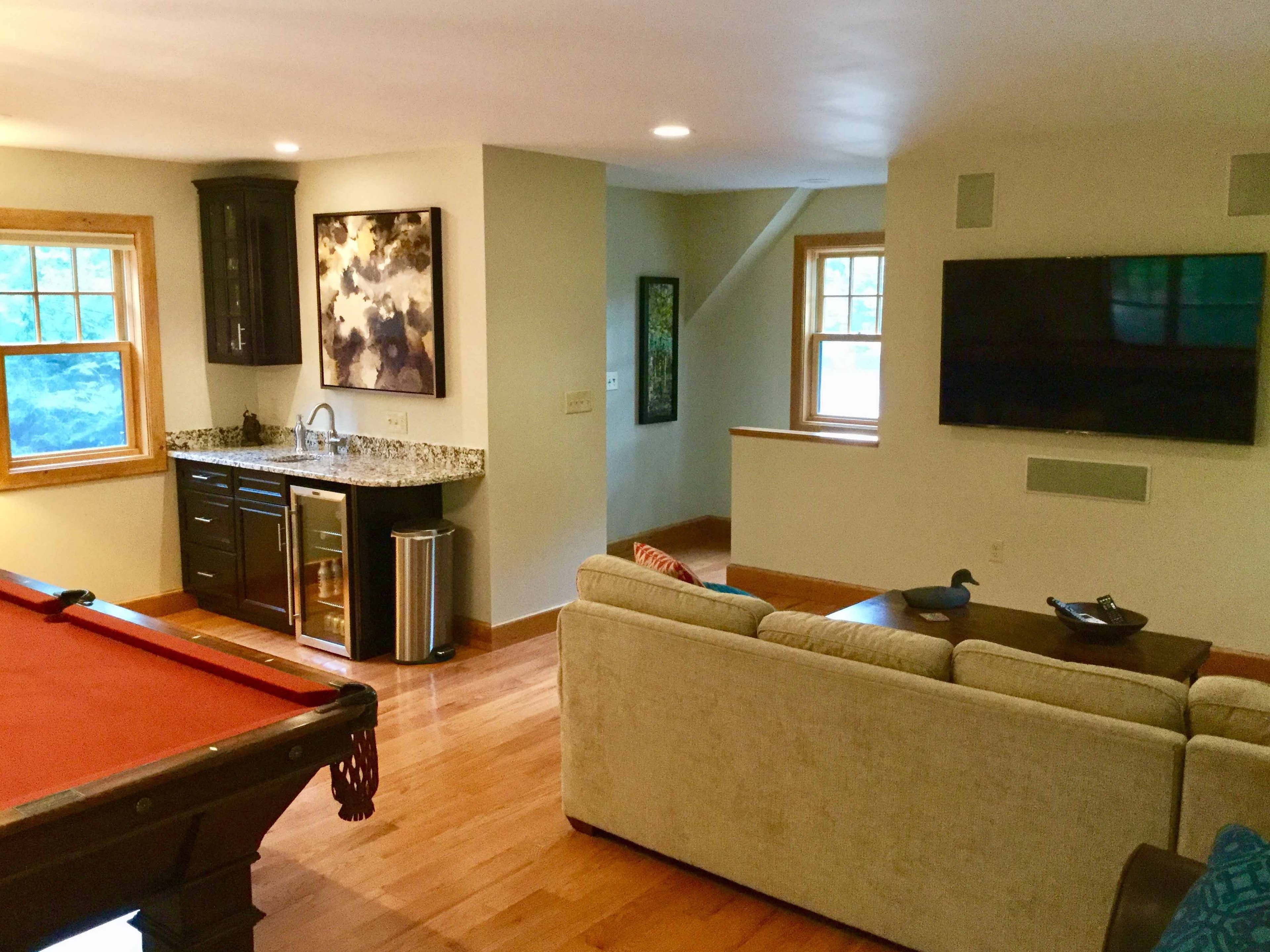 Finished bonus room showing wet bar with dark espresso cabinets, granite countertop, beverage fridge, and wall-mounted TV with surround sound speakers