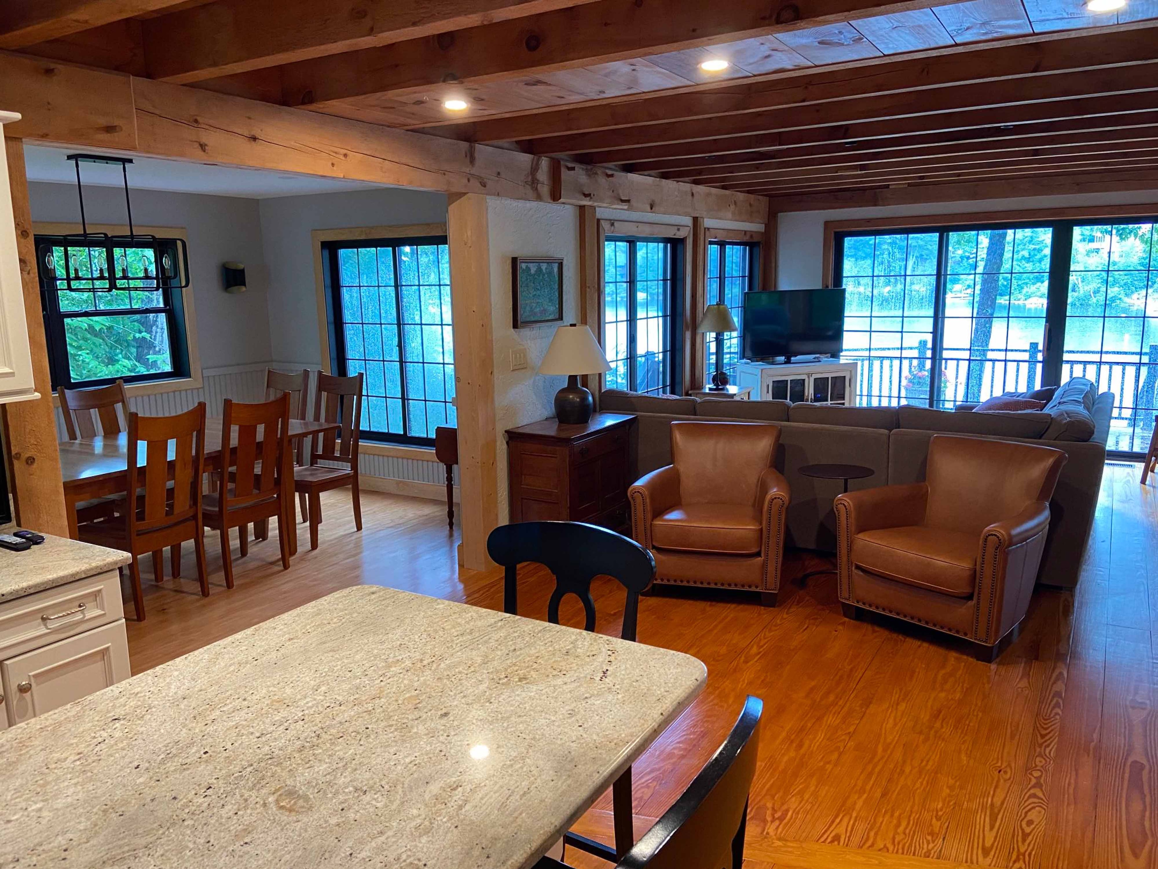 Open-plan lakeside living and dining room with timber beam ceiling, granite kitchen island, leather club chairs, and panoramic lake views through black-framed windows