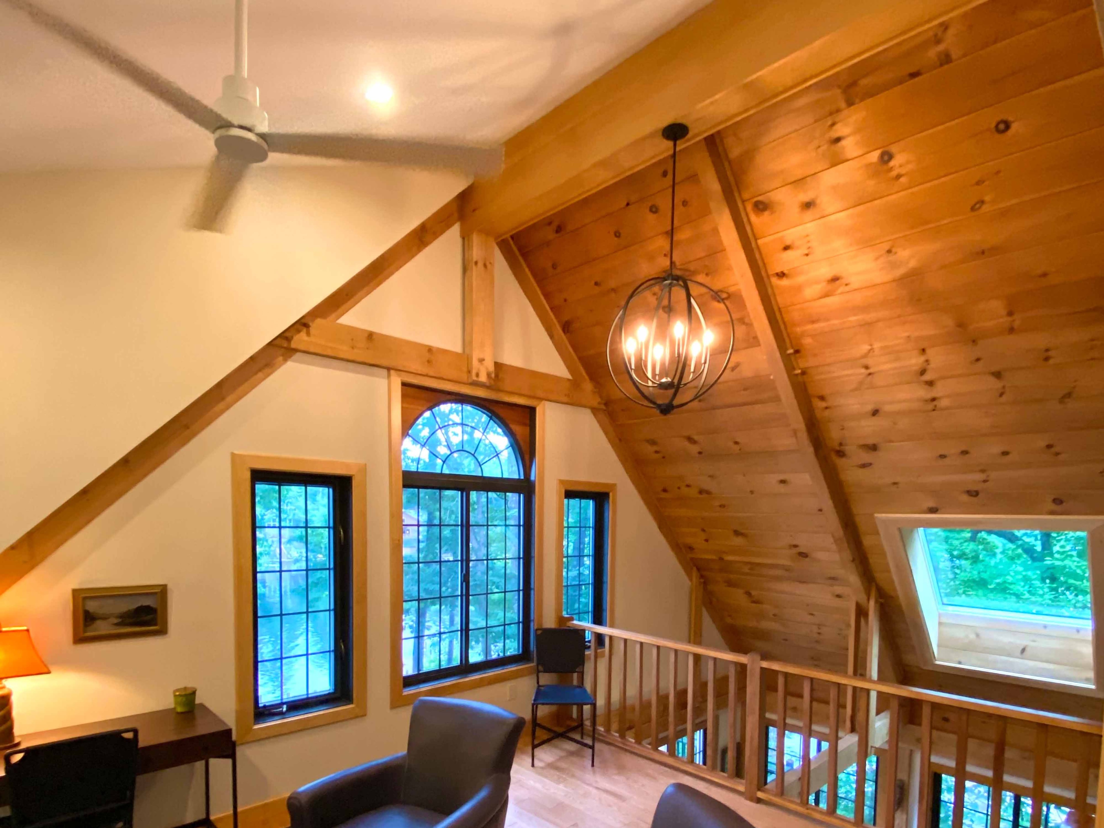 Dramatic timber-frame loft with knotty pine tongue-and-groove ceiling, exposed knee braces, arched Palladian window, orb chandelier, and hardwood flooring