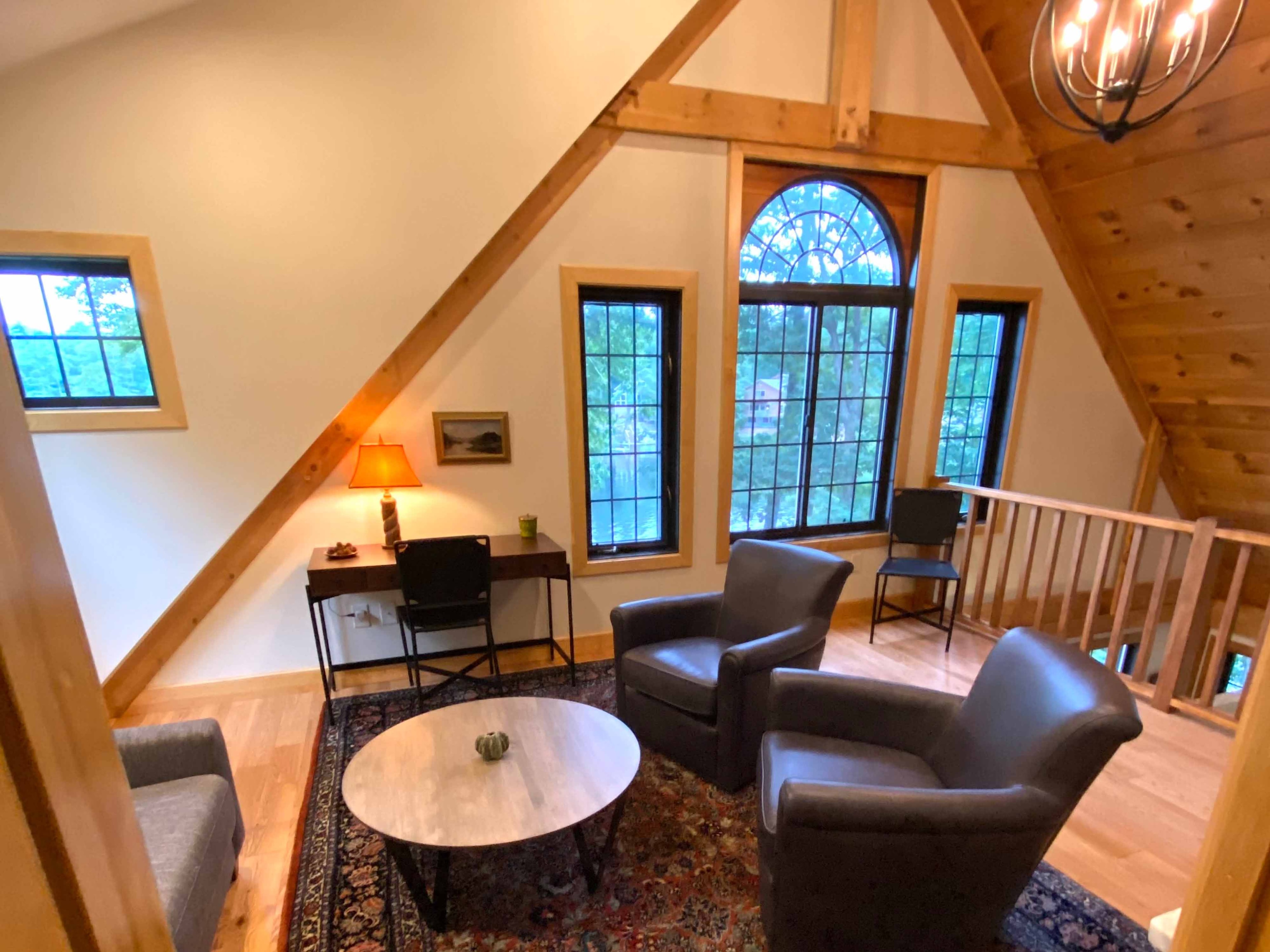 Loft sitting area viewed from above showing leather club chairs, Persian rug, round coffee table, arched window, and timber-frame knee braces
