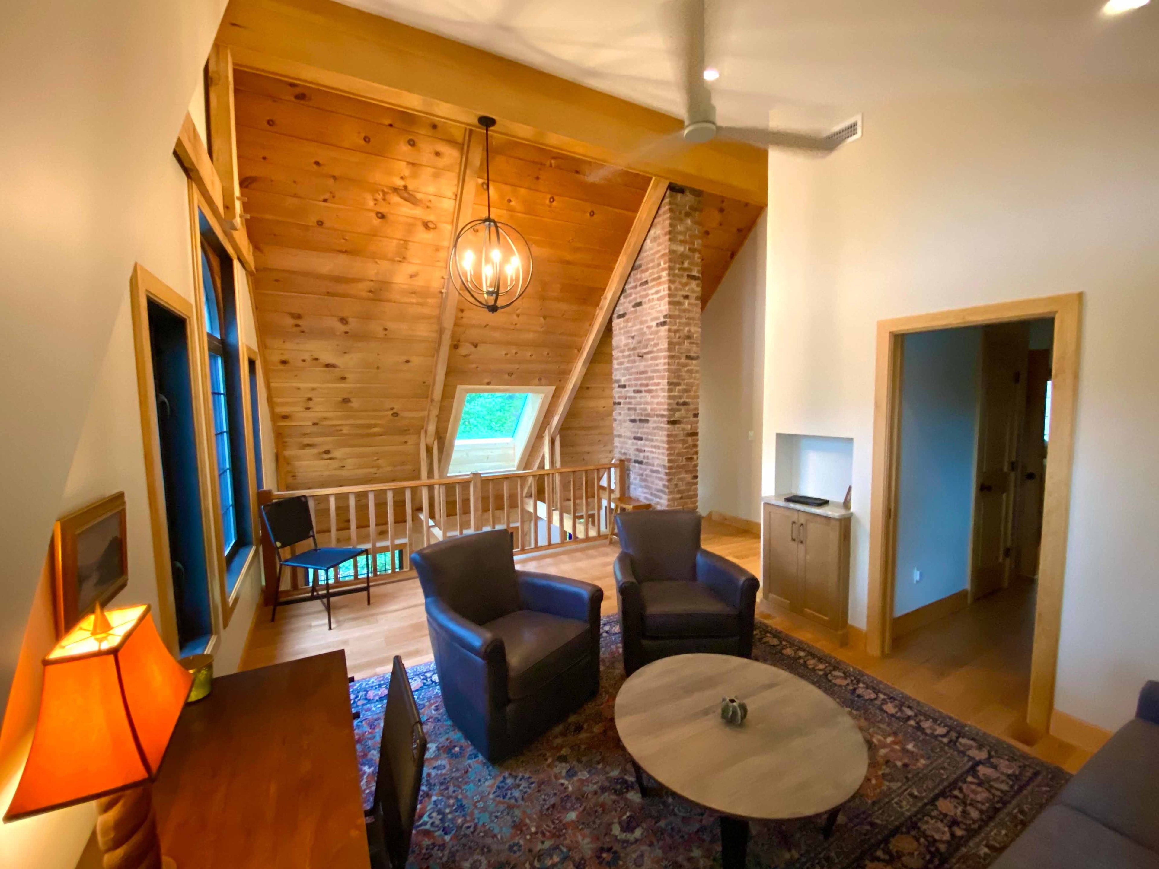 Loft sitting room with knotty pine cathedral ceiling, exposed timber knee braces, brick chimney column, leather club chairs, Persian rug, and orb chandelier