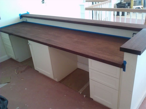 Custom built-in desk with rich dark walnut butcher-block countertop, white shaker base cabinets with drawers, and blue painter's tape on edges