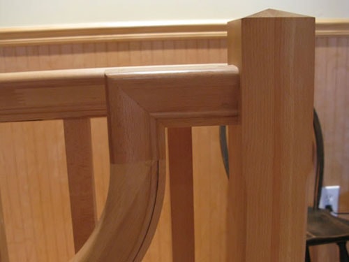 Close-up detail of oak stair railing joinery showing curved bracket, mitered cap rail, and square newel post with beadboard panel