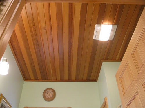 Cedar tongue-and-groove ceiling with recessed lighting and exhaust fan, viewed from stairwell with natural wood trim door and wall clock