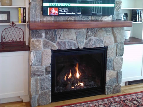 Completed fieldstone fireplace with walnut mantel shelf, wall-mounted TV, and flanking white built-in bookcases with hardwood flooring and Persian rug