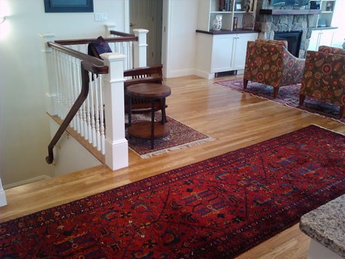 Open living room with white oak hardwood flooring, white baluster stair railing, stone fireplace with walnut mantel, and layered Persian rugs