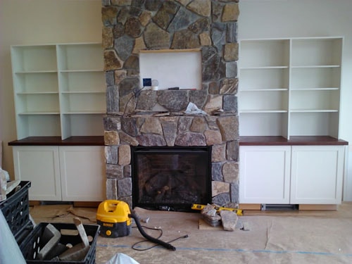 Fieldstone fireplace surround with flanking white built-in bookcases and dark walnut countertops under construction with shop vac and tools