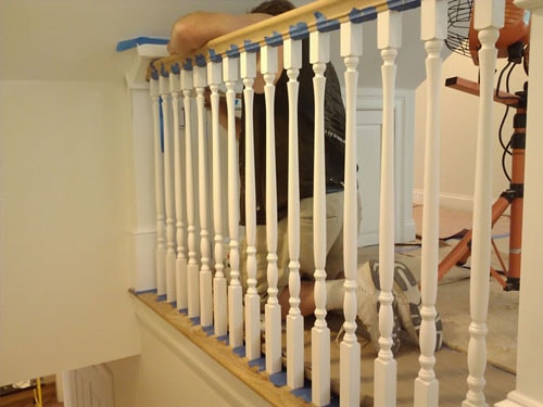 Craftsman installing white turned balusters on oak stair railing with blue painter's tape, fan and ladder visible in background