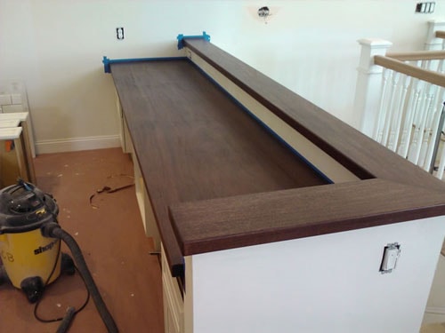 Dark walnut butcher-block bar countertop being fitted on white shaker cabinetry near stair railing opening with shop vac and tools