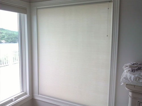 Cellular honeycomb window shade installed in bay window corner with lake view and white trim, pedestal table in foreground