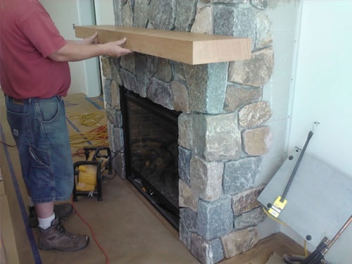 Craftsman holding walnut mantel shelf against completed fieldstone fireplace surround during installation with tools and extension cord on floor