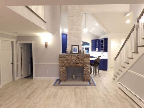 Dramatic open-plan remodel with freestanding stone chimney column, cobalt blue kitchen cabinetry, vaulted beadboard ceiling with pendants, white oak staircase, and wood-look tile floor