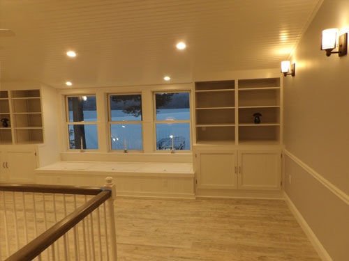 Upper loft room with full-width built-in window seat, flanking white bookcases with cabinet bases, beadboard ceiling, recessed lighting, and frozen lake view