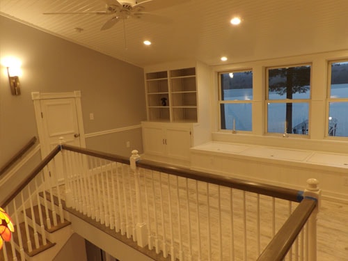 Loft landing with dark oak railing and white balusters looking toward built-in bookcases, window seat, and triple lake-view windows at dusk
