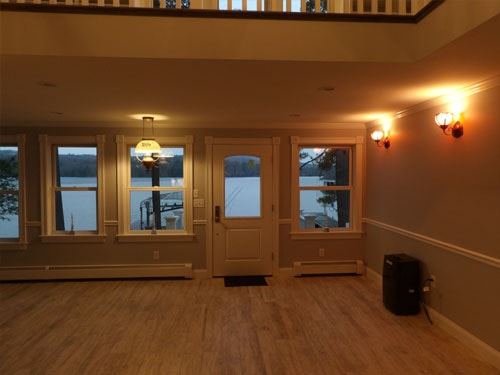 Finished lower-level entry with lake-view windows, white six-panel door, LVP flooring, wainscoting, and wall sconces at dusk