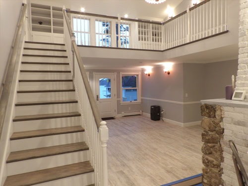 Full two-story view of straight-run staircase with white balusters, loft railing, lower entry with lake-view door, and stone chimney column
