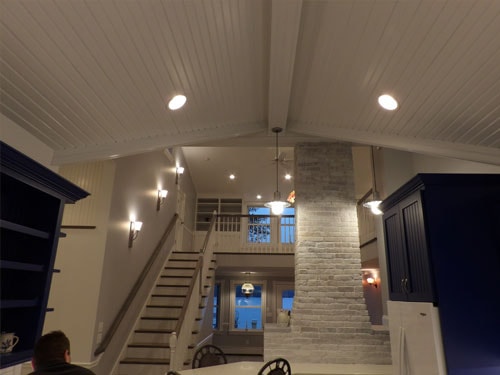 Vaulted white beadboard ceiling with exposed timber ridge beam, recessed lighting, wall sconces, stone chimney column, and staircase viewed from kitchen level at dusk