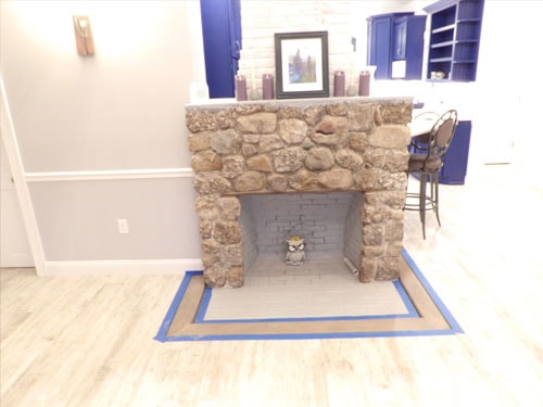 Completed fieldstone fireplace surround with cobblestone face, brick firebox, and blue painter's tape marking hearth border on new LVP flooring