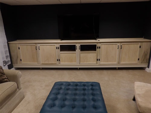 Full-width custom maple media cabinet with raised panel doors, black hardware, open center bay for AV equipment, and wall-mounted flat-screen TV