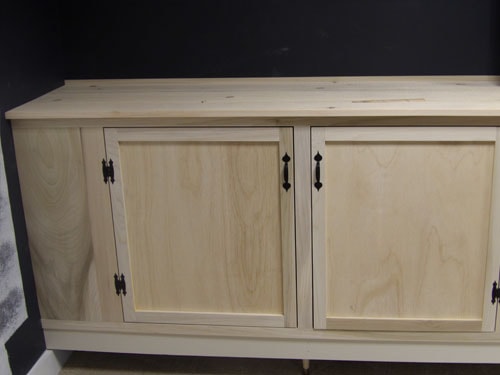 Close-up of left section of custom maple media cabinet showing raised panel doors, black bin-pull hardware, and butcher-block top against dark accent wall