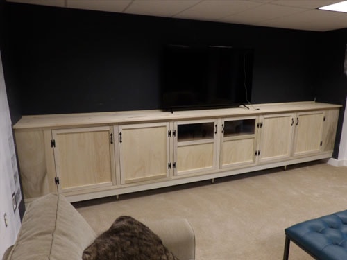 Full-width custom maple media cabinet from slightly wider angle showing all cabinet doors, open AV bay, wall-mounted TV, and tufted blue ottoman in foreground