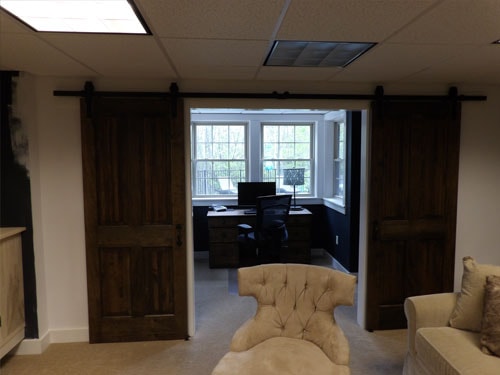 Custom double barn doors slid open revealing finished home office with bay window, built-in desk, computer setup, and tufted chaise lounge in foreground