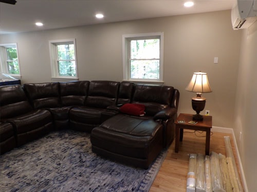 Finished basement living area with dark leather sectional recliner sofa, blue area rug, recessed lighting, two double-hung windows, and wall-mounted mini-split AC unit