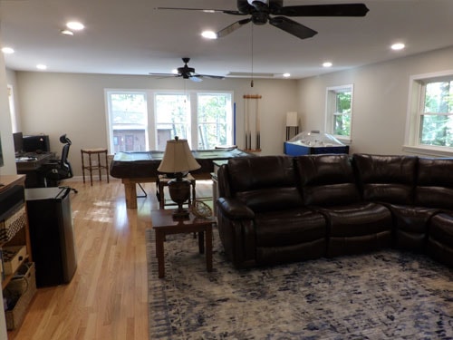 Wide view of finished rec room addition with pool table, air hockey table, dark leather sectional, blue rug, oak hardwood flooring, two ceiling fans, and multiple windows