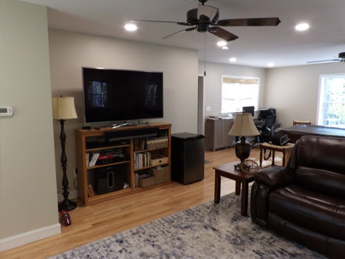 Finished rec room TV wall with large flat-screen, oak media console with shelving, dark leather armchair, blue rug, ceiling fan, and view into home office beyond