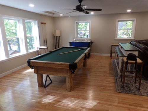 Finished rec room showing pool table with green felt, air hockey table, bar-height table with stools, ceiling fan, and multiple windows with wooded views
