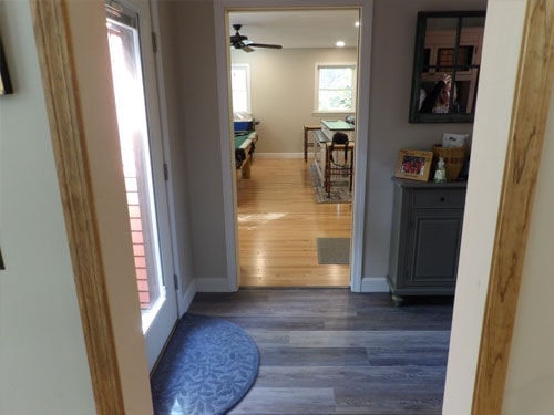 Mudroom-to-rec-room transition showing LVP flooring in entry and oak hardwood beyond doorway, with blue accent rug and natural wood door trim