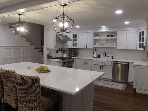 Stunning white shaker kitchen remodel with quartz waterfall island, farmhouse sink, subway tile backsplash, lantern pendants, wainscoting staircase wall, and dark hardwood flooring