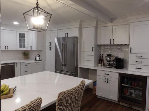 White shaker kitchen showing refrigerator wall with French door fridge, beverage fridge, coffee station, glass-front upper cabinets, and quartz island with wicker stools