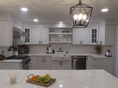 Straight-on view of white shaker kitchen sink wall with farmhouse sink, open shelving, subway tile backsplash, glass-front uppers, lantern pendant, and quartz island in foreground