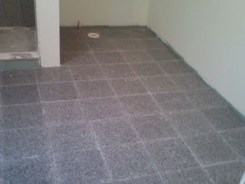 Gray granite tile flooring installation in progress in empty room with drain rough-in