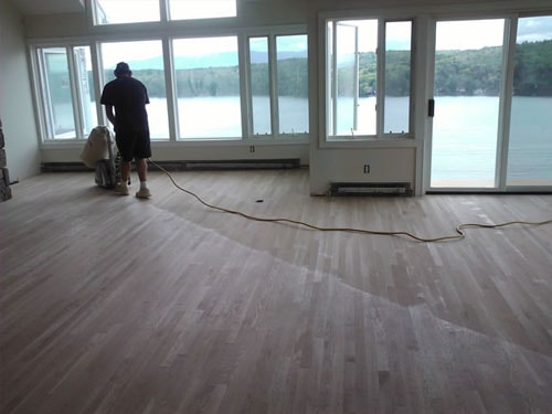 Worker sanding hardwood floors with floor sander in lakeside room with panoramic windows